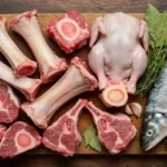 "A variety of raw soup bones, including beef marrow, chicken carcasses, lamb shanks, and fish heads, displayed on a wooden board with fresh herbs."