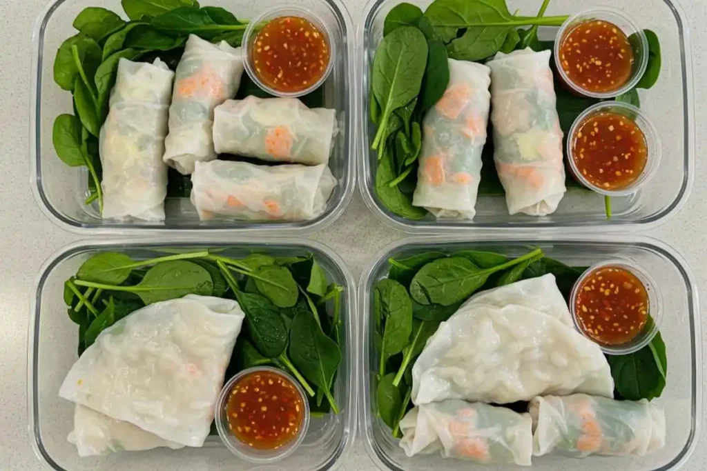  Neatly packed rice paper rolls in meal prep containers with fresh greens and sauce.

