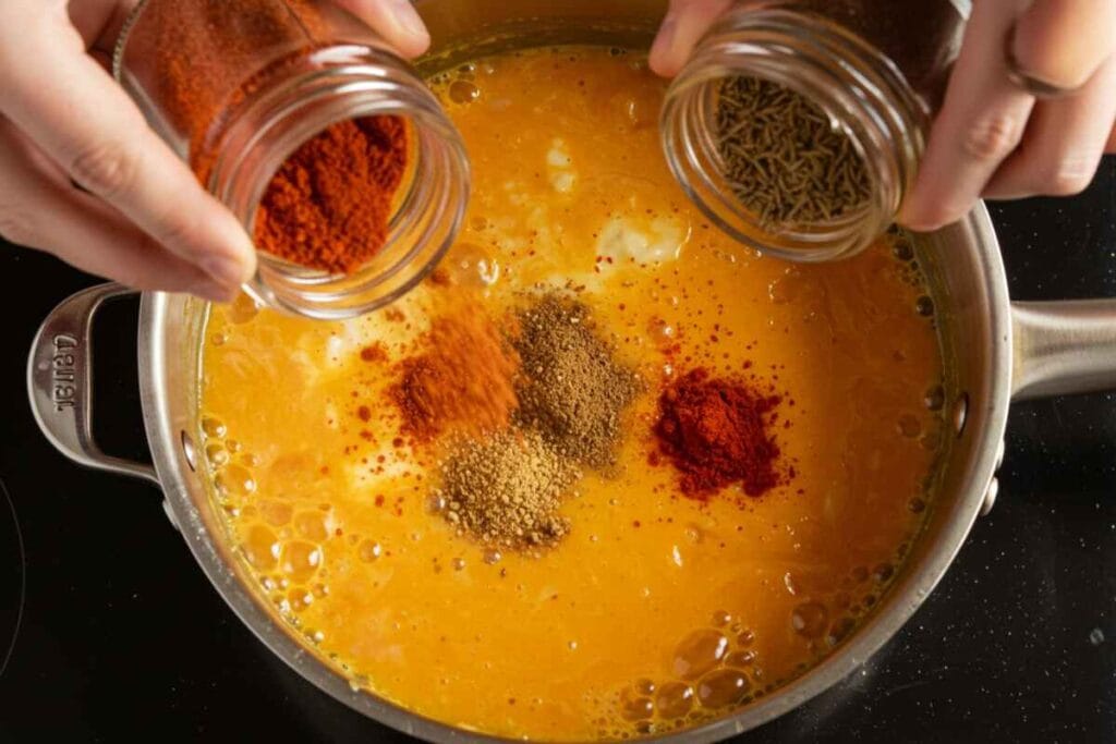 A variety of taco spices being sprinkled into a saucepan of cheese sauce